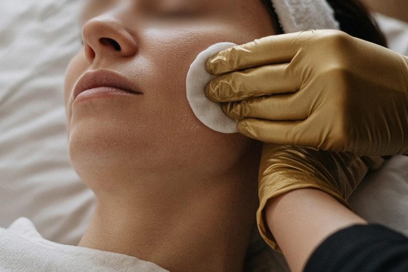 Gold-gloved hand cleansing a client's cheek with a cotton pad